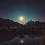 A peaceful view of moonlit mountains reflected in a tranquil lake under a starry sky.