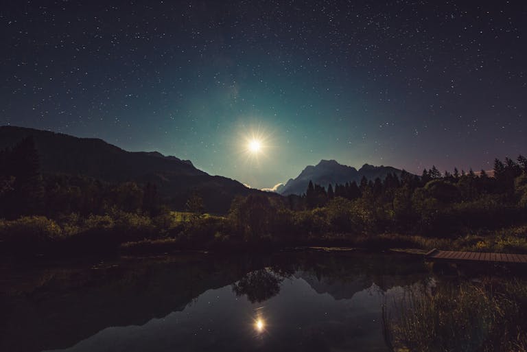 A peaceful view of moonlit mountains reflected in a tranquil lake under a starry sky.