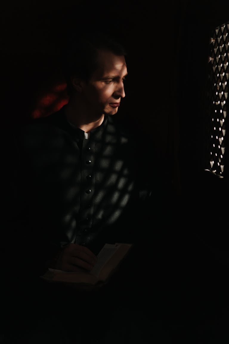 A priest sits in a dim confessional holding a Bible, reflecting faith and spirituality.